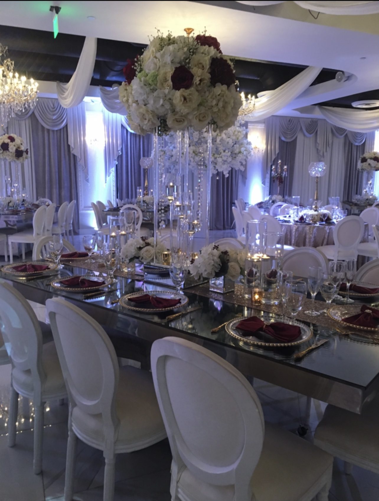 Elegant banquet hall with white chairs, mirrored tables, gold-rimmed plates, burgundy napkins, and tall centerpieces of white and red flowers; draped curtains and chandeliers add to the luxurious atmosphere.