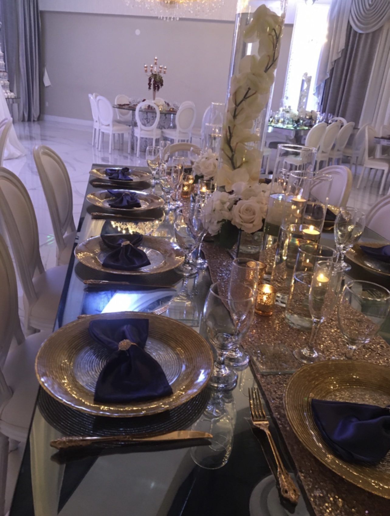 A formal dining table is set with gold-rimmed plates, navy blue napkins, gold flatware, glassware, candles, and elegant white flower centerpieces in a luxurious banquet hall with white chairs and drapes.